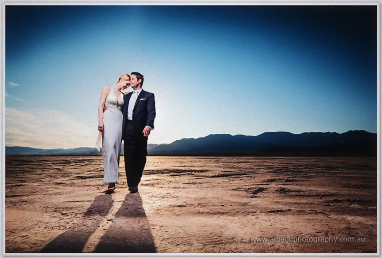Bride and groom walk close with heads together, long shadows stretching across the playa