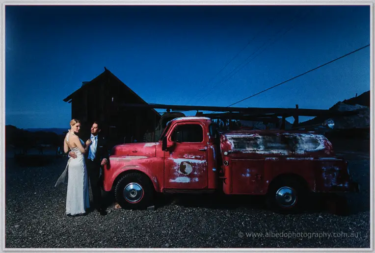 Bride and groom stand by a weathered red tanker truck at a desert homestead at dusk