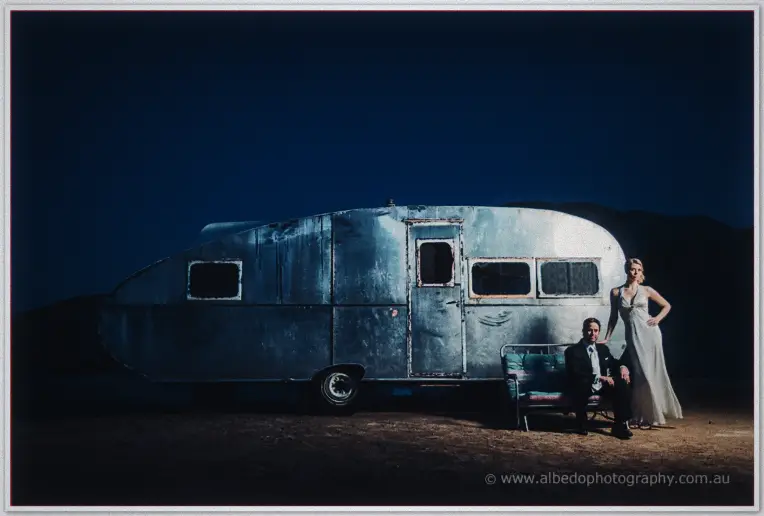 Groom sits on a bench with his bride beside a vintage silver trailer under a deep blue dusk sky