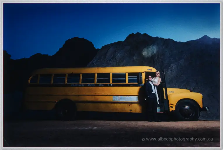 Bride and groom pose at the door of a faded yellow school bus beneath dark desert hills
