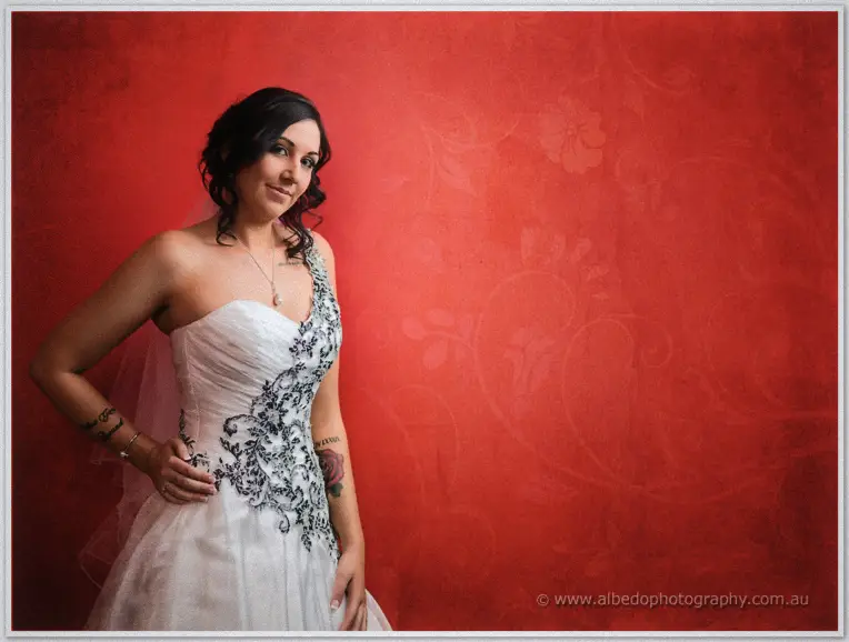 Jenna poses one hand on hip before a textured red wall, tattoos tracing her arm beneath soft light