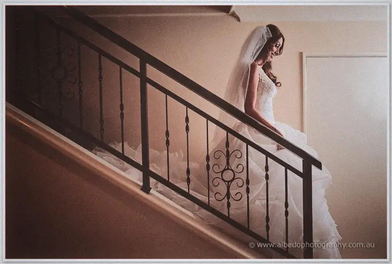 Bride in a ruffled gown and veil glancing back over ornate black stair railings in warm light