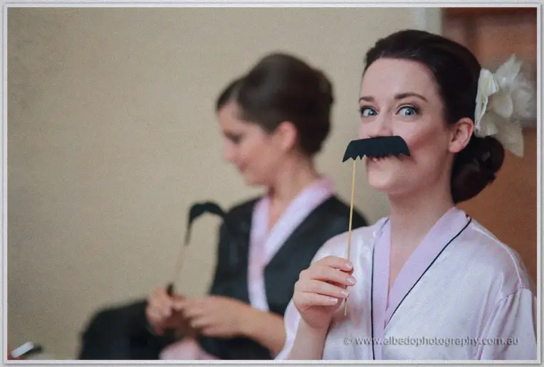 Bridesmaid in pink robe pulling a funny face with a paper moustache prop