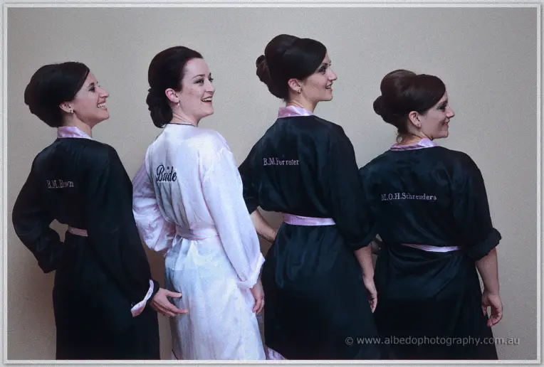 Bride in white robe glancing back between three bridesmaids in personalised black robes