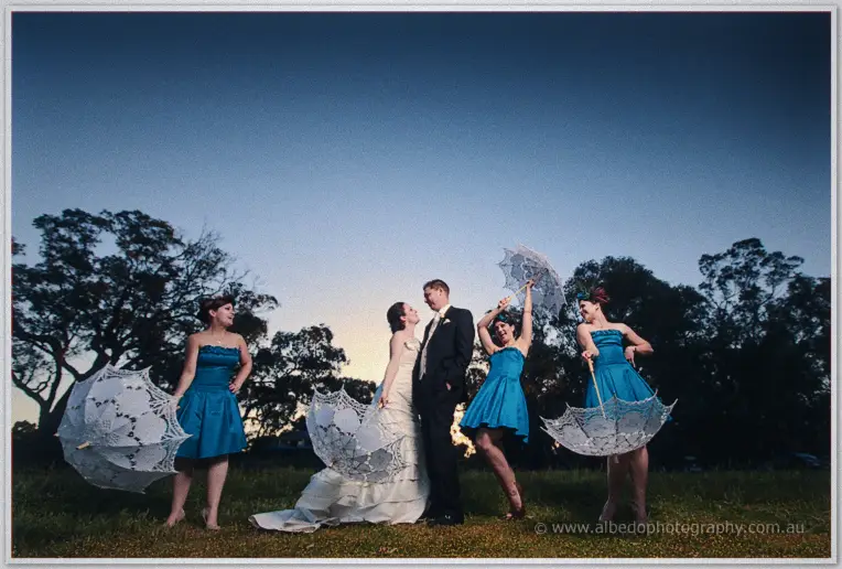 Phil kisses Felicity at dusk while four teal-dressed bridesmaids twirl lace parasols around them
