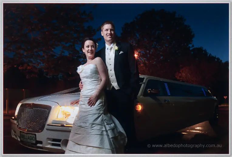 Felicity laughs beside Phil in front of the headlit stretch limousine under the trees after dark