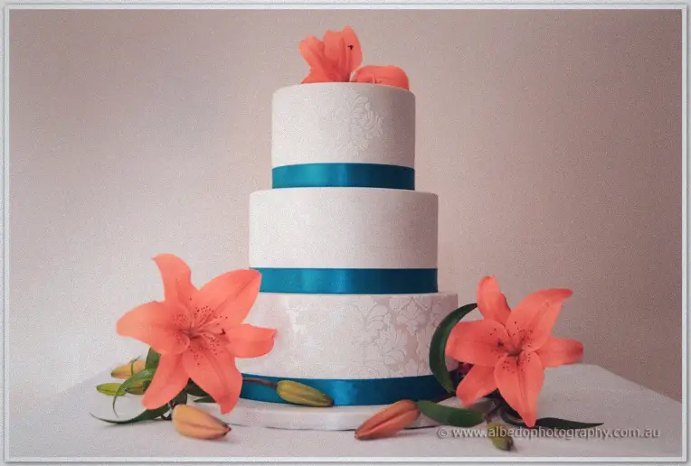 A three-tier white cake wrapped in teal ribbon and crowned with coral lilies awaits the cutting