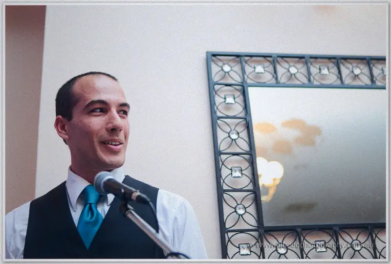A groomsman in a teal tie grins into the microphone during a lively reception speech