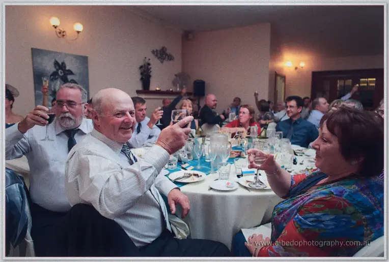 Smiling wedding guests raise their wine glasses in a toast across the warmly lit tables