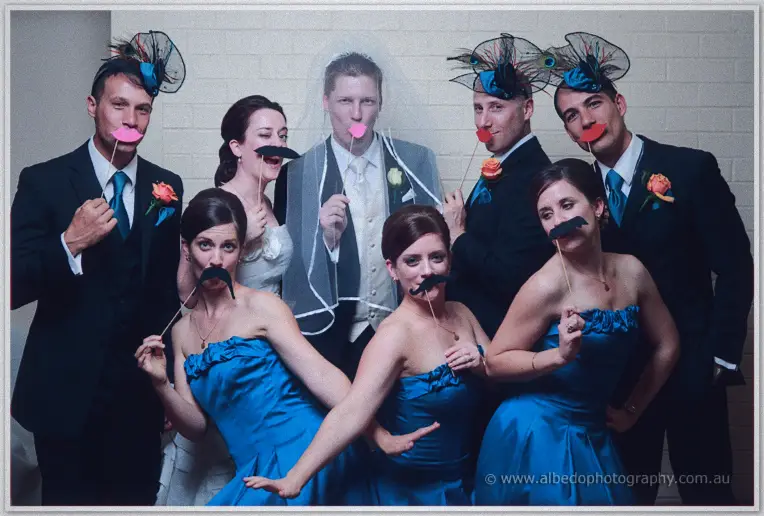 The bridal party in blue gowns and dark suits poses with moustache props in the photo booth