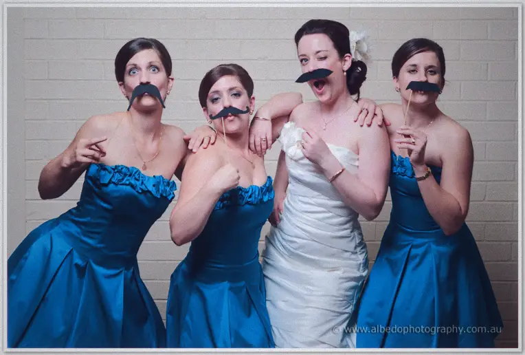 Felicity and her bridesmaids in strapless blue gowns grin behind moustache props in the booth