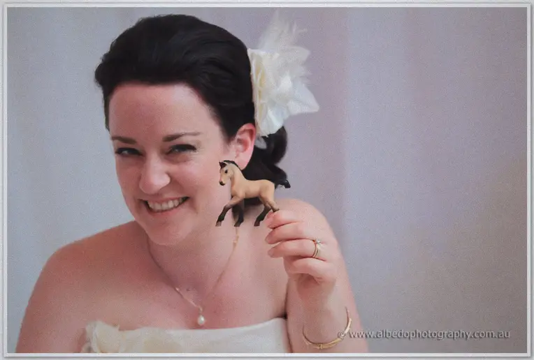 A laughing Felicity in her white feather fascinator holds up a tiny toy horse in close-up
