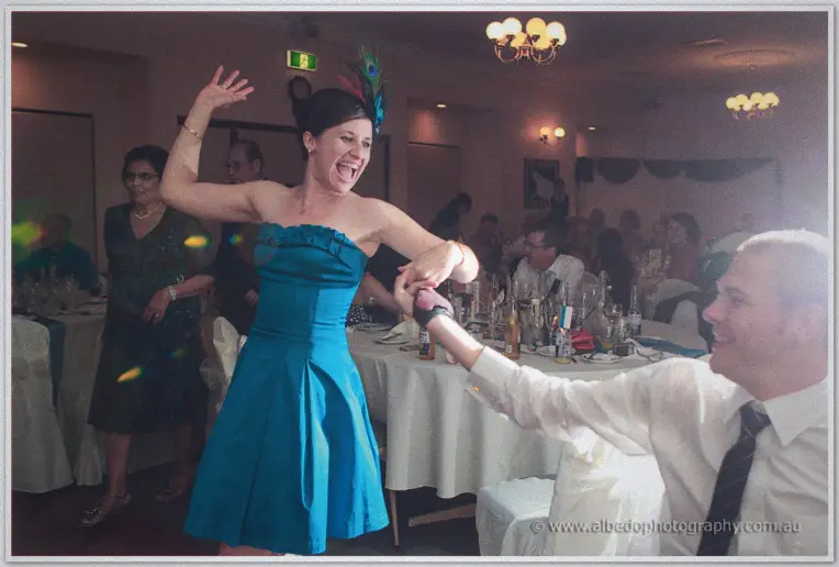 A woman in a teal dress and peacock fascinator dances with arms raised among the guests