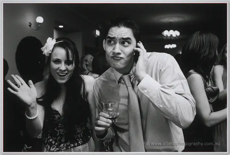 A grinning guest pulls a playful face, drink in hand, beside a woman in a flower headband