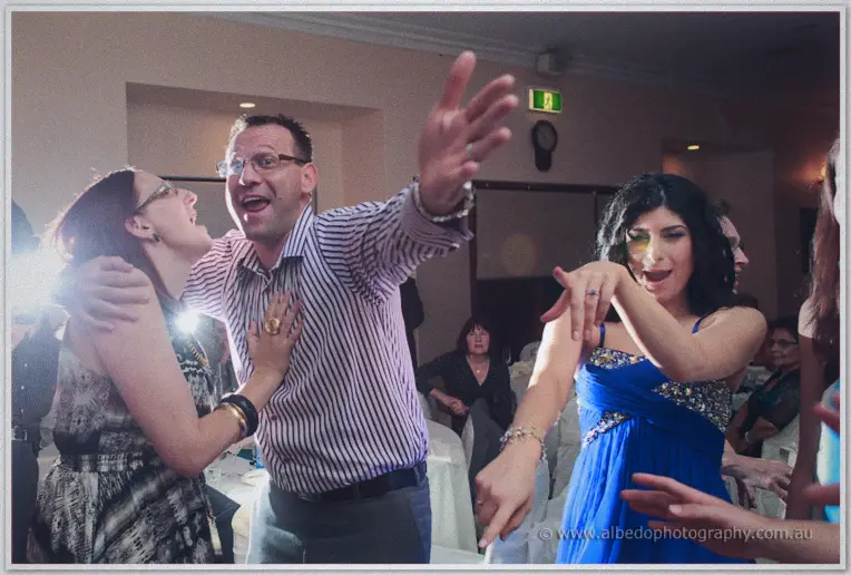 A man in a striped shirt sings with his arm outstretched between two dancing women in blue