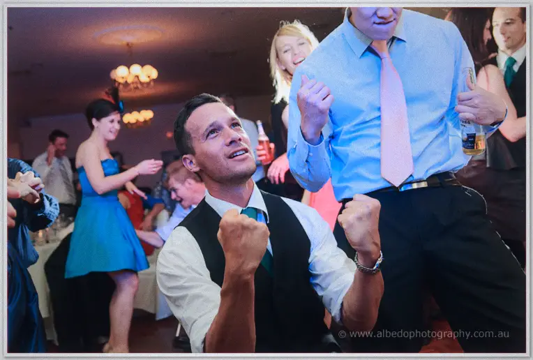 A groomsman in a teal tie pumps both fists on the dance floor as the party rages behind