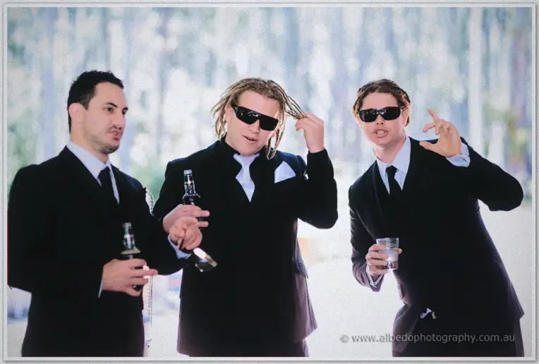 Andrew and his groomsmen joke in sunglasses with beer bottles against a bright forest backdrop
