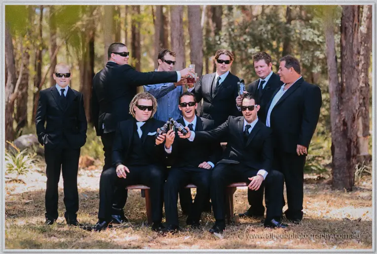 The groomsmen raise beer bottles in a group toast under tall trees in matching black suits