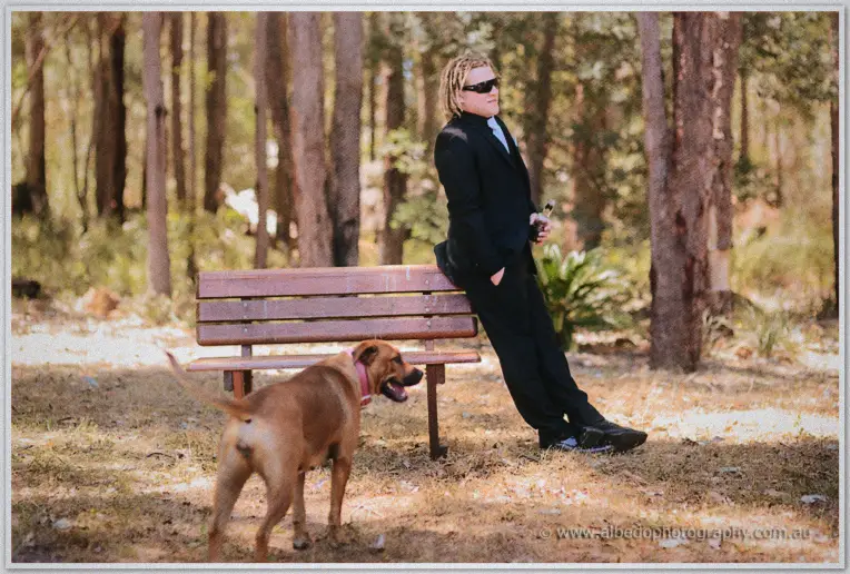 Andrew leans on a park bench with a drink as a dog wanders past in the sunlit bushland