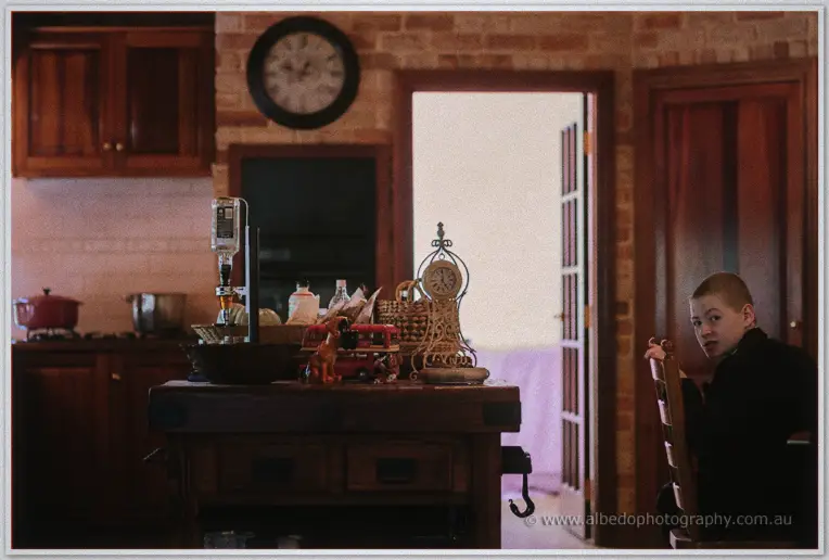 A young page boy in a dark suit peeks over a timber chair in a warm brick kitchen