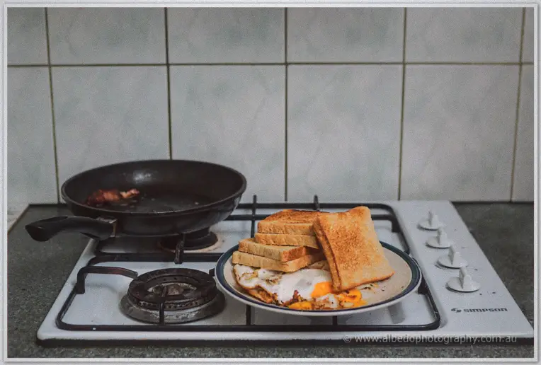 A plate of eggs and toast sits beside a frying pan on a tiled stove at breakfast