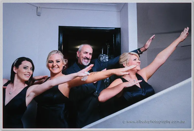 Bridesmaids in navy dresses strike playful poses on a staircase beside a smiling older man