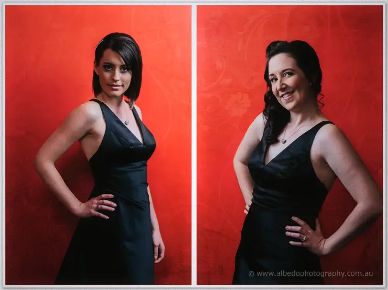 Two bridesmaids rest their hands on their hips in navy satin gowns before a bold red wall