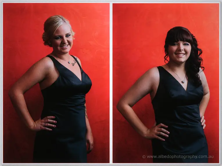 A blonde and a dark-haired bridesmaid grin side by side in navy dresses against the red backdrop