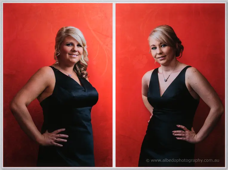 Two smiling bridesmaids in navy gowns stand shoulder to shoulder beside the deep red wall