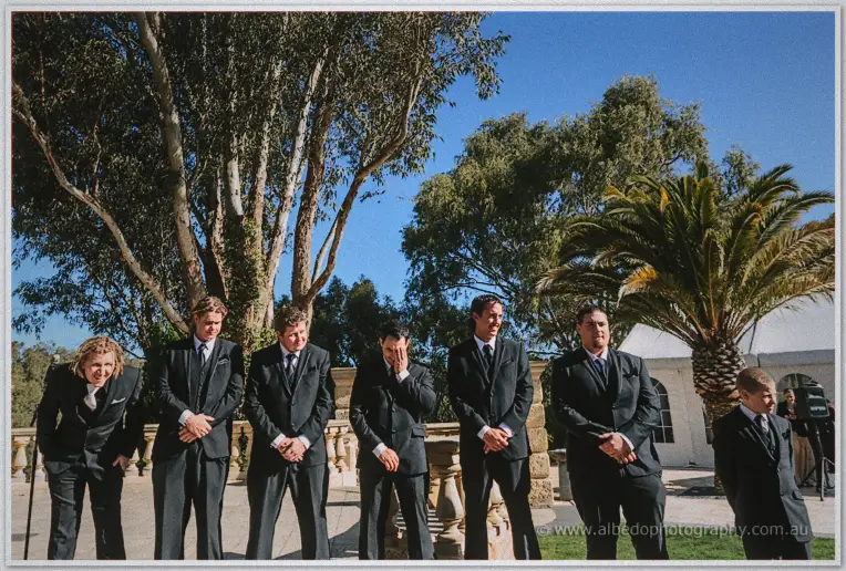 Groomsmen in black suits wait and laugh beneath gum trees on a sunny terrace