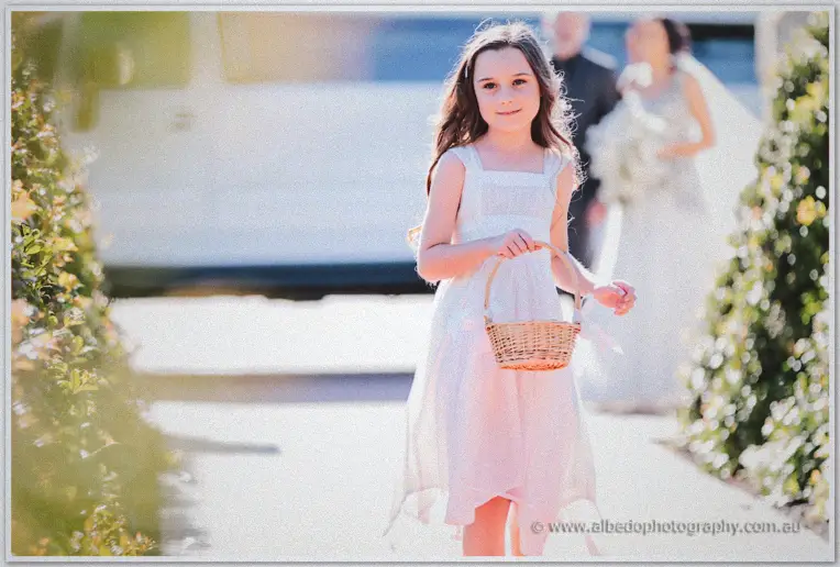 A young flower girl in pink carries a wicker basket down the path as the bridal party follows behind