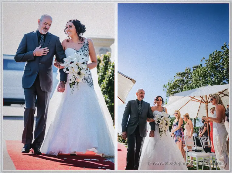 Father and bride walk the red carpet together with guests watching from white chairs in the sun