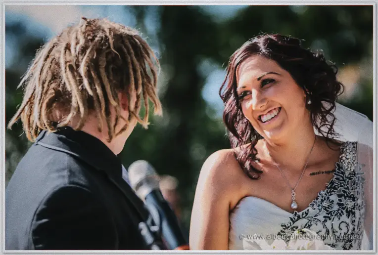 Jenna laughs during the ceremony as Andrew faces her before a microphone in dappled shade