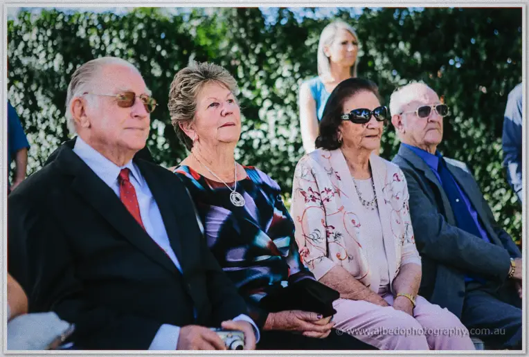 Older guests in sunglasses watch the ceremony from the front row against a leafy hedge