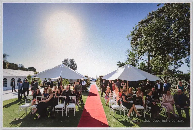 White chairs line a red carpet aisle between parasols as guests settle before the ceremony