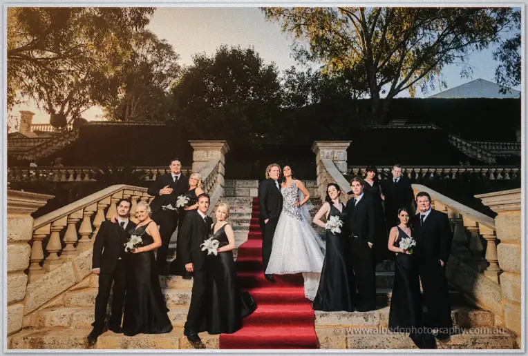 Bridal party in black gowns and suits flank Jenna and Andrew on a red-carpeted staircase at golden hour.