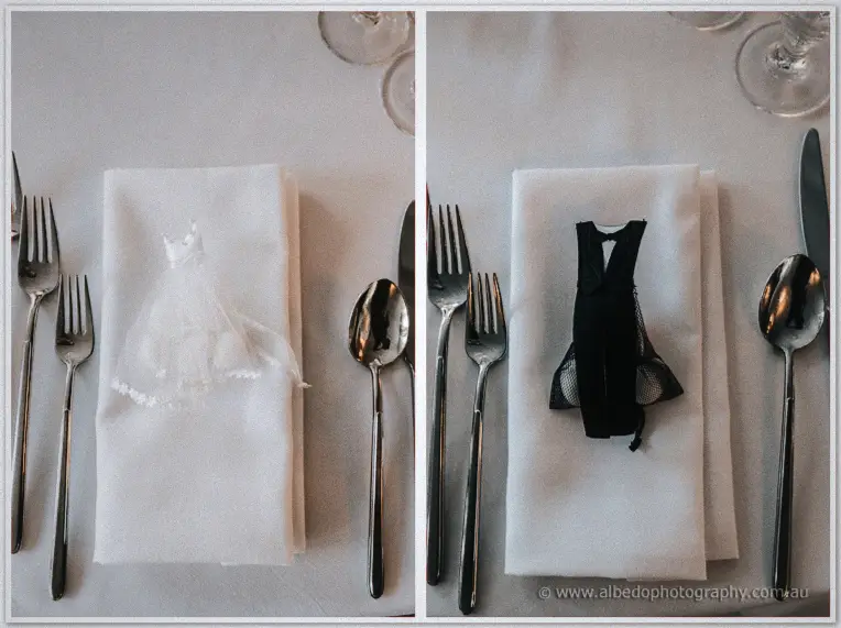 Place settings show miniature bride and groom outfits folded onto white napkins beside silver cutlery.