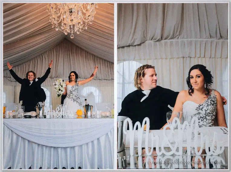 Jenna and Andrew fling their arms wide at the bridal table beneath a crystal chandelier.