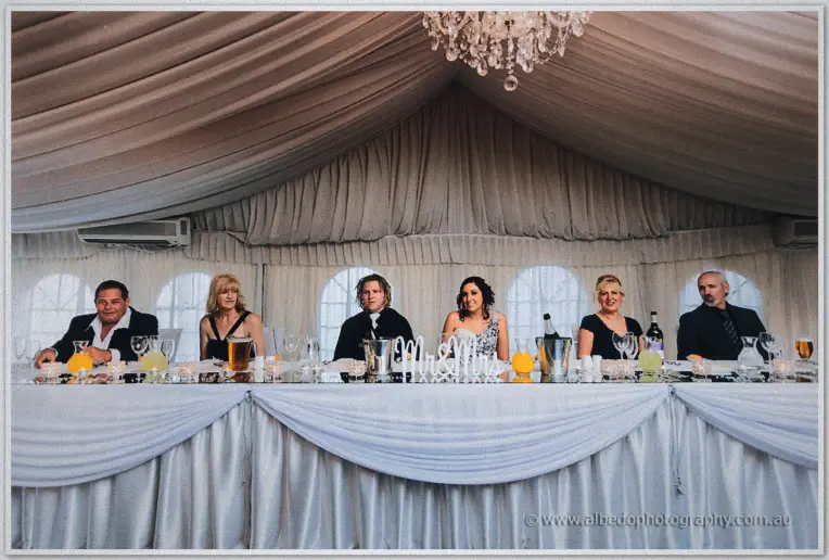 The full bridal table sits under the marquee chandelier, Jenna and Andrew centred behind a Mr and Mrs sign.