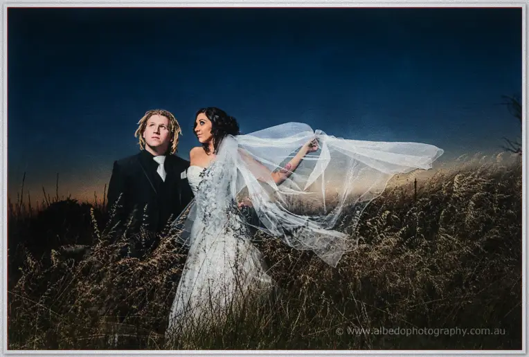 Jenna and Andrew stand in windswept grass at dusk as her veil billows under a deepening blue sky