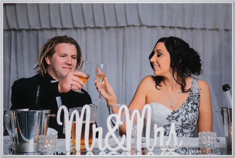 Andrew and Jenna toast with champagne flutes at the bridal table behind a script Mr and Mrs sign.