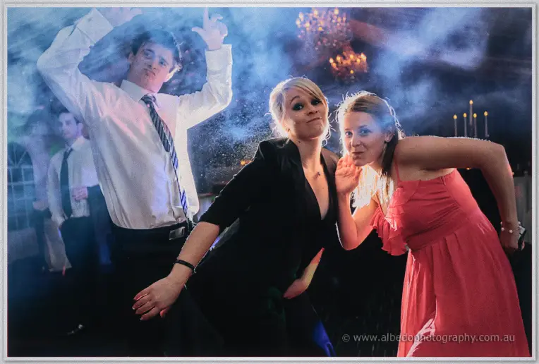 Three party guests in shirt and tie, black and coral dresses pull funny poses in the smoke under chandeliers.
