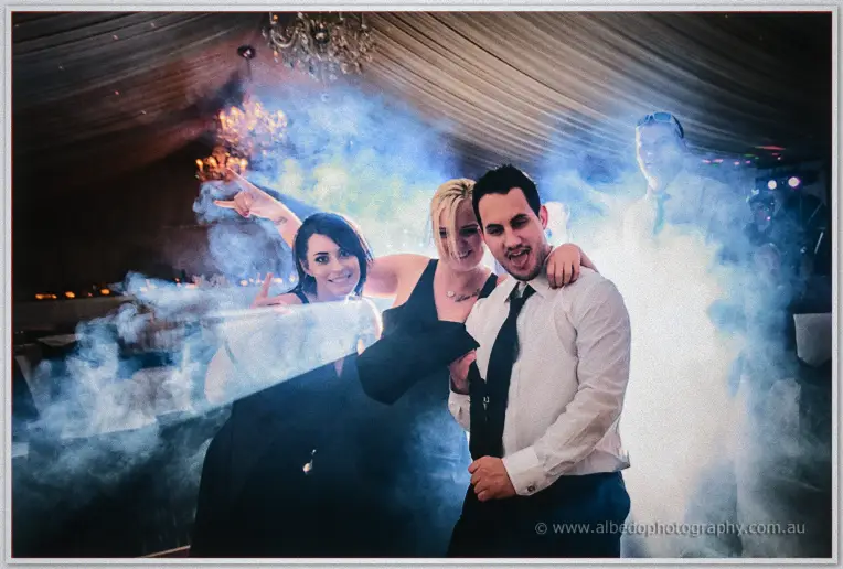 Two women in black and a man in white shirt and tie point and grin through the fog on the dance floor.