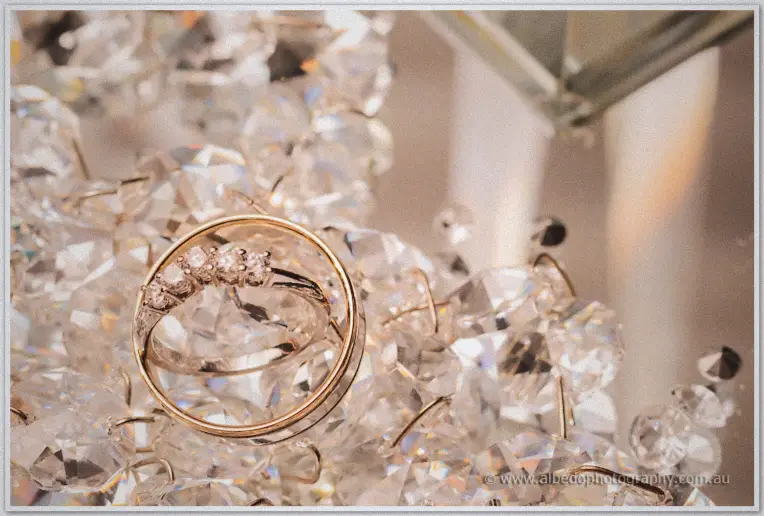 Gold wedding bands and a diamond-set engagement ring nestled in sparkling crystal chandelier droplets.