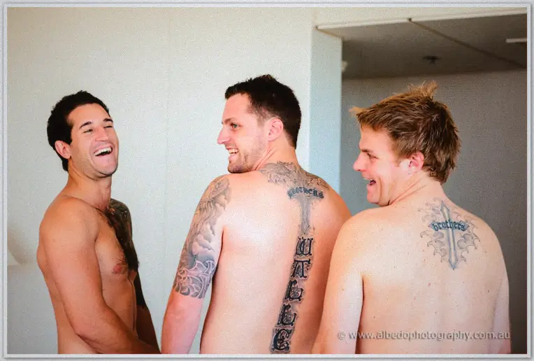 Three shirtless groomsmen laugh together, matching brotherhood tattoos across their backs