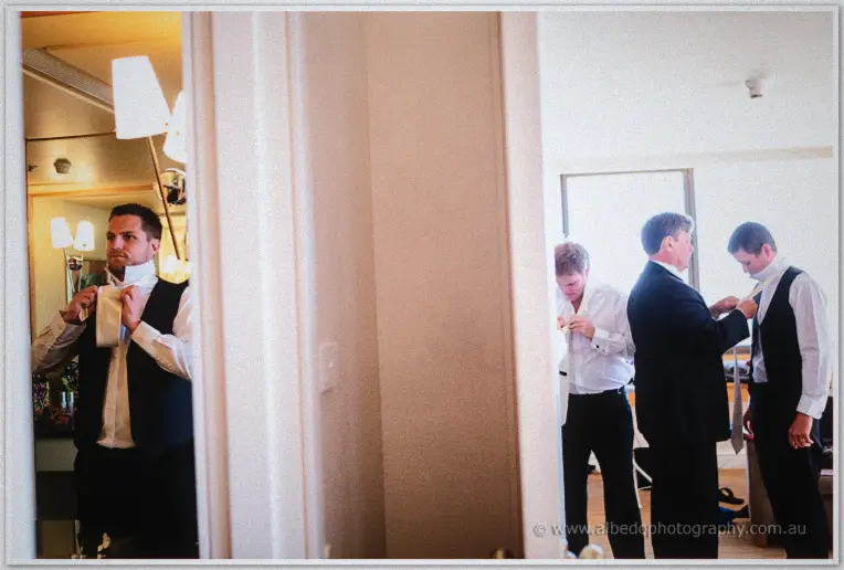 Groomsmen fasten ties and vests around a hotel mirror as Nathan readies his collar
