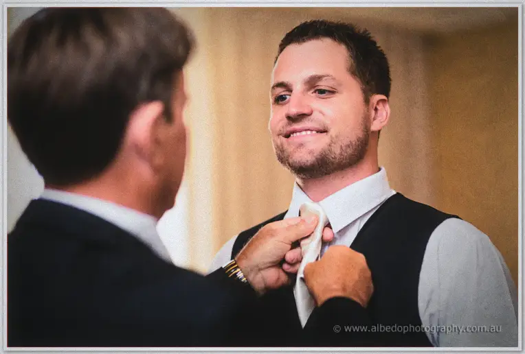 His father ties Nathan's cream necktie as the groom smiles in warm morning light