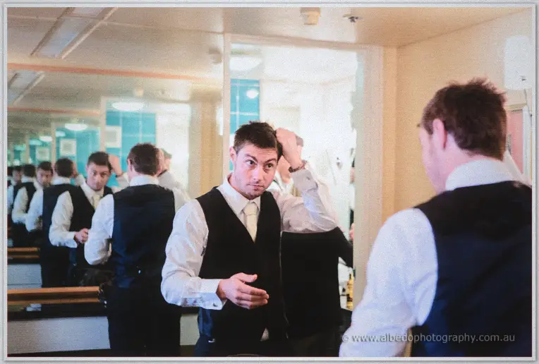 A groomsman fusses with his hair in the mirror while others adjust vests behind him