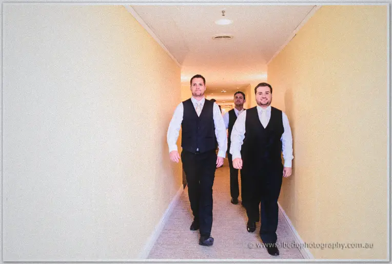 Groomsmen stride down a cream hotel hallway towards the ceremony