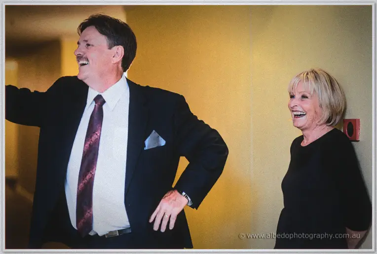 A suited father and a blonde guest in black share a laugh in the hotel's golden hallway